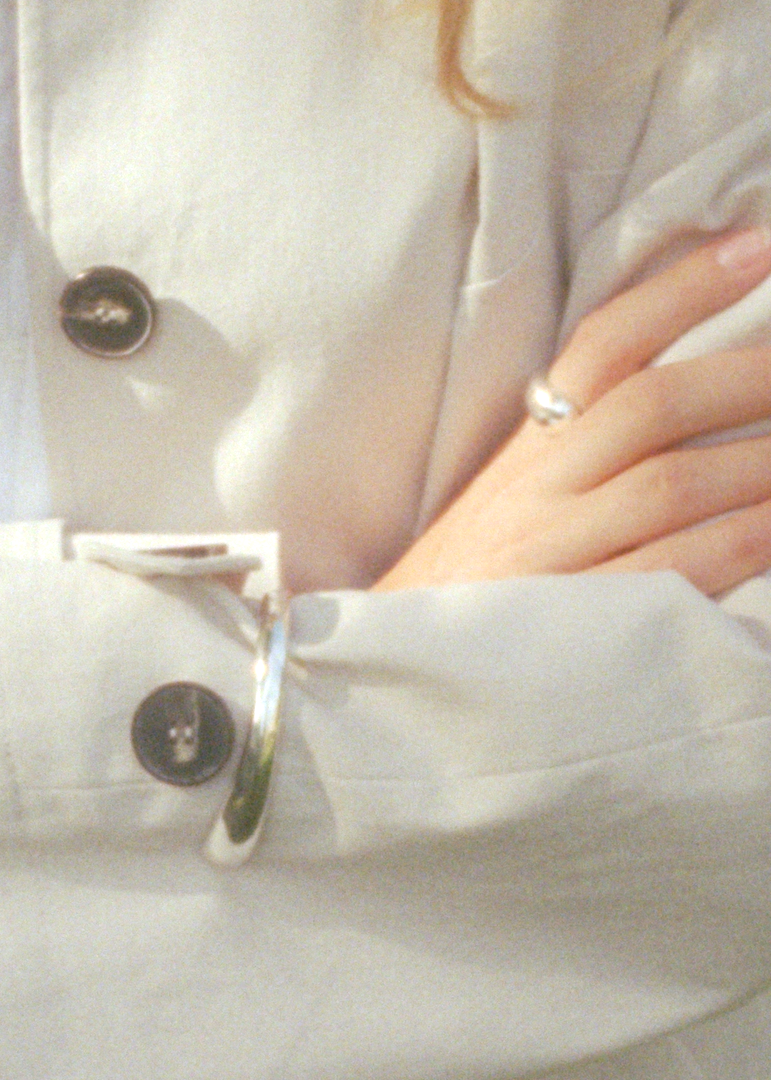 A close-up of a person outdoors wearing a light-colored coat with dark buttons, arms crossed. The person is wearing silver rings on both hands, and a silver bracelet is tucked into the coat pocket.